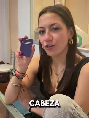 A woman with dark hair and gold hoop earrings holds a blue card with text in a light-colored room. The text 'CABEZA' is overlaid at the bottom of the frame. She has multiple bracelets on her wrist, and a necklace.