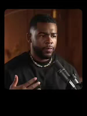 A medium shot of a man with a dark complexion, wearing a black top and a diamond necklace, is speaking into a microphone. The background is a muted brown, suggesting an interior setting. The man is the central figure, with his focused expression and hand gestures indicating he is actively engaged in the conversation. The lighting is soft and diffused, highlighting his features.