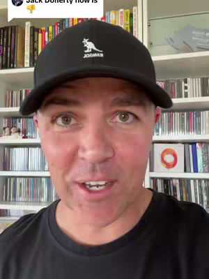A man is centered in a close-up shot, wearing a black cap and a black t-shirt. Behind him, a bookcase is filled with books and CDs. He is looking directly at the camera with his mouth slightly open.
