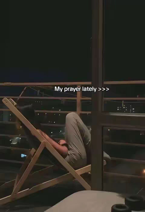 A person is lying on a wooden folding chair on a balcony at night. The background is dark, with city lights faintly visible. The person is wearing casual clothes and has their legs elevated while leaning back. Text overlaid on the image says, "My prayer lately >>>".