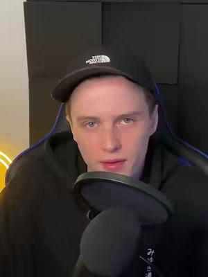 A man wearing a black cap and hoodie is looking at the camera while holding a microphone. The background is dark with a geometric pattern.