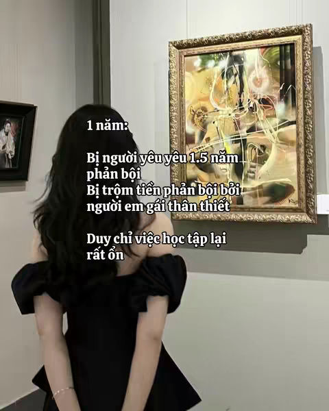 The cover image presents a young woman from the back, slightly turned to the left, in front of an art gallery wall. She is wearing a black off-the-shoulder dress. To the right side of her head, a framed painting with abstract shapes and colors hangs on the wall, and on the left, a dark framed picture. Text overlay, in simple white font, is positioned to the left, describing a personal horoscope, in Vietnamese.. The overall composition has a soft, muted color palette with focus on the figure and text.