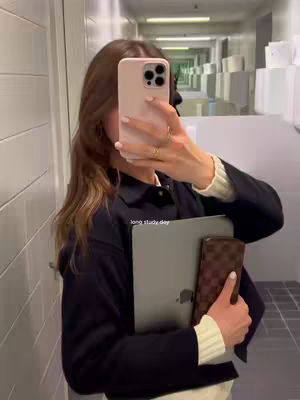 A woman is taking a selfie in a brightly lit hallway, holding a phone up to her face with her left hand. The phone is in a pink case, obscuring most of her face, but her hair is visible, and she is wearing a dark jacket over a cream-colored top. She's carrying a laptop and a wallet. The hallway has white paneled walls, a series of square cutouts, and a reflective ceiling.