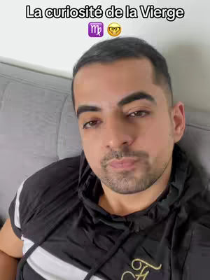 A man with short dark hair and a beard looks towards the camera. He's wearing a black hoodie with white stripes on the sleeves and a gray couch is in the background. Above his head is the text 'La curiosité de la Vierge' and a Virgo symbol.