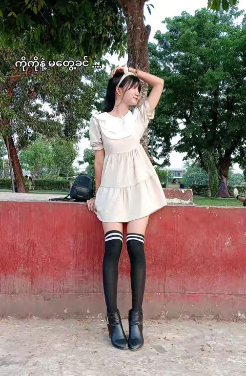 A woman in a light-colored dress is positioned in front of a vibrant red wall. She is wearing black knee-high socks with white stripes at the top, and black boots. The background shows foliage and trees suggesting an outdoor setting.