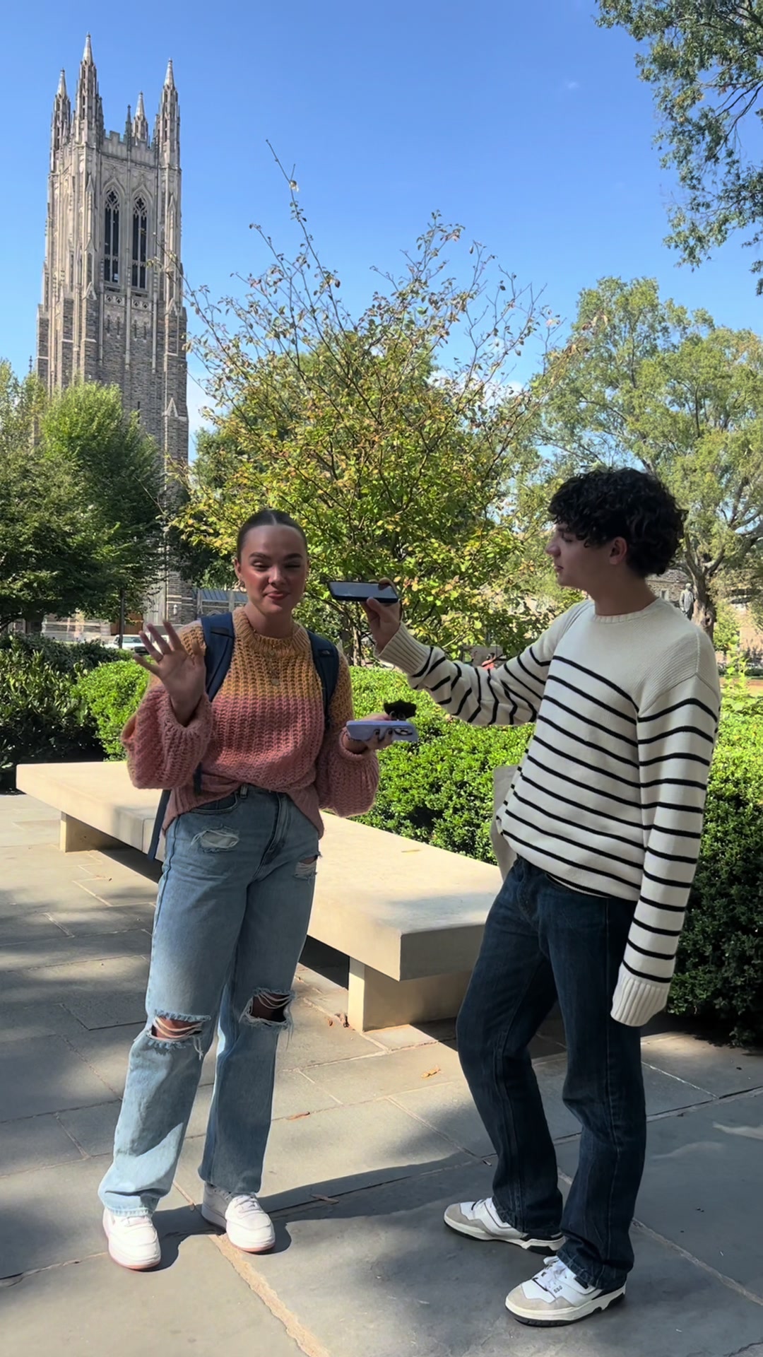 A sunny outdoor shot features two college students, one with a backpack, interacting in front of a tall university building. They appear to be filming, with one holding a phone and the other modeling an outfit consisting of a pink and yellow sweater and ripped jeans.