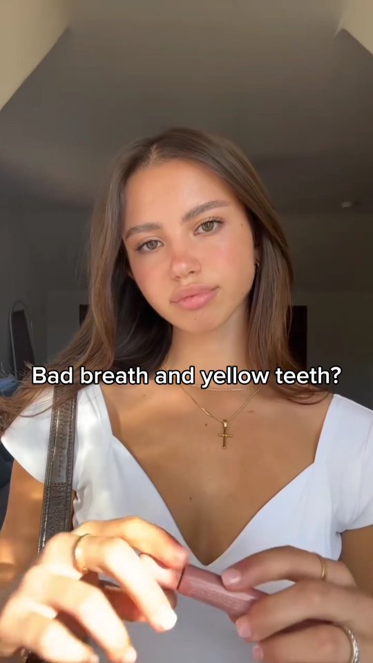 A young woman with fair skin, light brown hair, and a white top is holding a tube of lip gloss. The text overlay reads, "Bad breath and yellow teeth?"