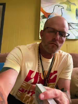 A man with glasses is holding a deck of cards. The background includes yellow wooden wall, a painting with a bird and some part of the white wall.