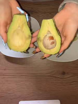 A top-down shot featuring two hands, each holding a half of an avocado on a small white plate. The avocado halves are vibrantly green. The background includes a wooden table, and the hands have ornate nail art.