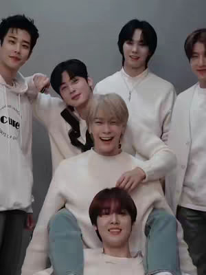 A group of six young men pose closely together, smiling at the camera. The men are dressed in shades of white and cream-colored sweaters and hoodies. The composition is from a medium angle with the individuals in a tight cluster. The background is a solid, neutral grey.