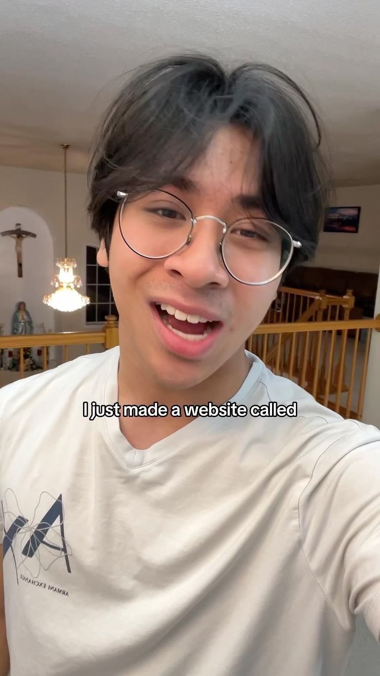 A young man with dark hair and glasses is smiling and talking. He's wearing a light-colored t-shirt in front of a home interior.  There is a cross on the wall in the background.