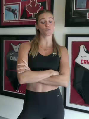 A woman with blonde hair, wearing a black sports bra and leggings, stands with her arms crossed. Behind her are framed pictures, some with a Canadian theme. She appears to be talking, with a slight pout.