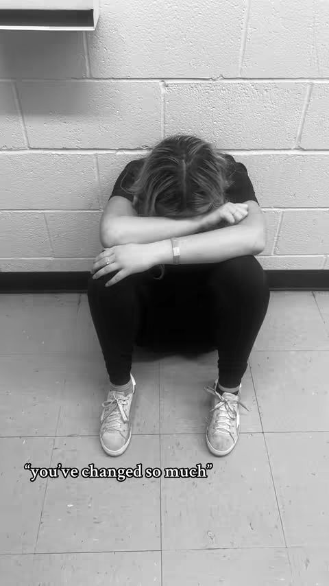 A grayscale image shows a person seated on the floor, with their knees drawn up toward their chest and their head resting on their arms, which are crossed. The person wears black clothing and sneakers. The background is a white brick wall and a gray tile floor. Text overlayed on the bottom reads "you've changed so much".