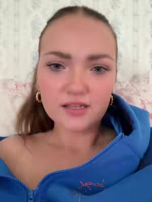A young woman is shown in a close-up shot, looking directly at the camera. She has fair skin, with light-colored eyes, and her hair is pulled back. She's wearing large gold hoop earrings and a blue hoodie, with a light-colored floral pattern visible behind her. The lighting seems natural and soft.