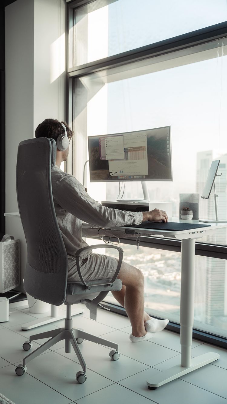 A man sits at a modern desk by a large window, working on a computer. He's wearing headphones and casual clothing. The scene is bright and airy, with views of city buildings visible outside the window.