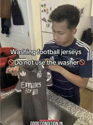 A man stands in a kitchen holding a football jersey over a sink. Text overlays indicate this is a tutorial on washing jerseys. The man is wearing a dark blue shirt with white stripes. The background shows a sink, cabinets, and a refrigerator.