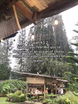 The image captures a scenic outdoor setting featuring a wooden structure with a thatched roof, which looks like a gazebo, surrounded by lush greenery. There are several people gathered at the structure. The backdrop is filled with tall, green trees, with soft lighting. The sky is overcast, with a few visible water droplets, creating a serene ambiance. A text overlay quotes a Bible verse.