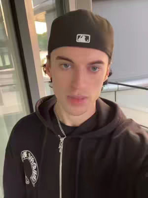 A young man wearing a black baseball cap and a black zip-up hoodie stands in front of a window. He is looking at the camera with a neutral expression. The background is softly lit, suggesting an indoor location.