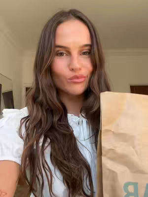 A young woman with long brown hair is holding a brown paper bag in front of her. She is smiling slightly, and the background suggests an indoor setting with neutral colors. The bag has some green text and the woman is wearing a white top.
