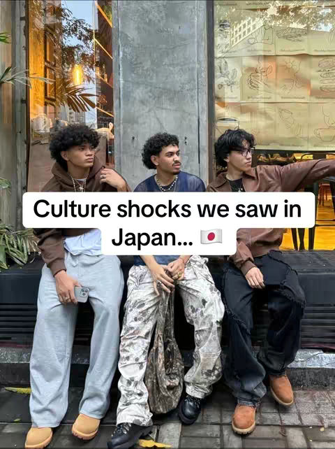 Three young men are seated on a low wall or bench, likely in an urban setting. The central figure is wearing patterned light-colored pants and a dark shirt holding a camera. Those on either side are in more casual attire. In the background is a glass window and text overlaid on a white rectangle that reads, 'Culture shocks we saw in Japan...'. A Japanese flag emoji is placed to the right of the text.