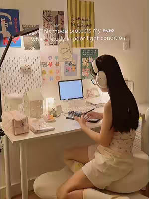 A well-lit room with a young woman seated at a desk, possibly studying. The desk is white and cluttered with items like a laptop, keyboard, and other stationery. The woman is turned away from the camera, wearing headphones and glasses, interacting with something on the desk. The background is a wall adorned with posters and decorations. There is text overlaid on the image.