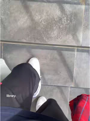 An overhead shot showing the lower body of a person standing on light gray, square tiles. The person is wearing a pair of black pants, white sneakers, and a navy blue shirt. A portion of a red and black patterned item is visible on the right side of the image. The overall scene is brightly lit.