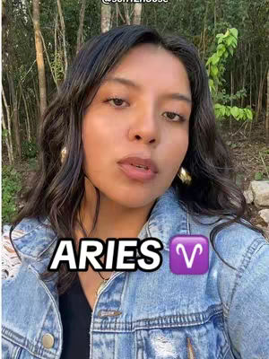 A selfie-style shot features a young woman with dark hair, a denim jacket, and gold hoop earrings. Overlayed on the image are the words "ARIES" in bold white font, with a purple Aries symbol next to it. The background suggests an outdoor setting with trees.