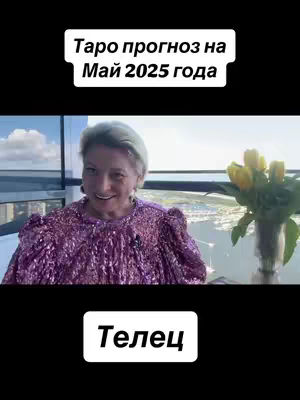The image shows a mature woman in a sparkly purple top, smiling. Above her is the text "Таро прогноз на Май 2025 года" in a white box. Below, the text "Телец" is also in a white box. In the background, a cityscape is partly visible through a window, with a vase of yellow tulips to the woman's right.