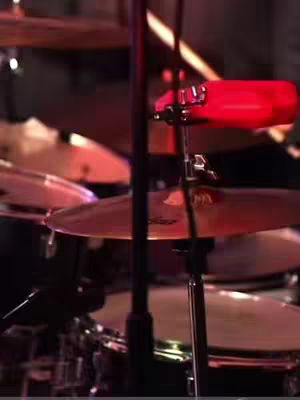 The image shows a close-up of a drum set. The focus is on the cymbals and a bright red percussion instrument attached to a stand. The cymbals are circular and metallic, with the drumsticks and parts of the drums visible. The lighting is dim, giving a slightly hazy appearance.