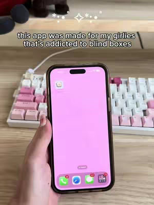 A hand holding a pink iPhone displaying an app, with a white and pink keyboard in the background. Text overlay says, 'this app was made for my girlies that's addicted to blind boxes'.