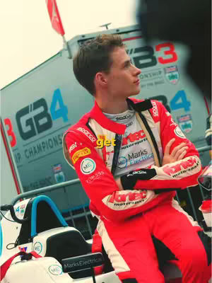 A young man in a red and white racing suit poses looking away from the camera. He is leaning against a race car, with race signage in the background.