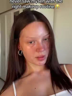 A woman with long brown hair is shown close-up, with one side of her face having no makeup on. She wears hoop earrings and a white top.