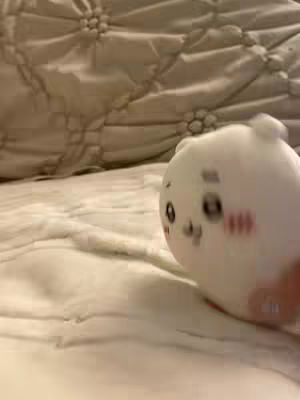 A plush toy sits on a white comforter. The toy is white, with eyes and red blush marks, and a hand is visible in the bottom right corner. The background is slightly out of focus.