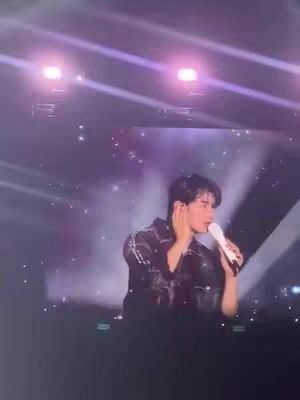 A man with black hair is on a stage, holding a microphone and singing. He appears to be performing in front of lights and a screen. The stage is dark, which casts the man in shadow.