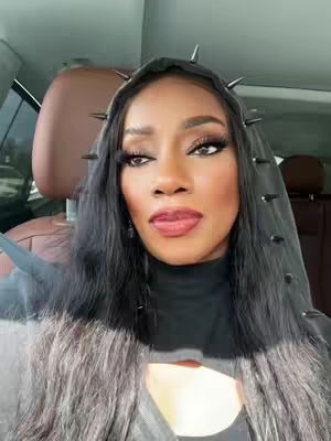 A woman with long black hair and a spiked hood is featured in a selfie. She is inside a vehicle, with the interior visible behind her. Her makeup is heavy and she is wearing a black top. The lighting appears to be natural with some shadows.