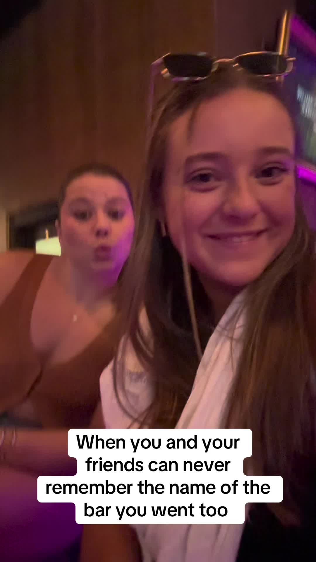 Two young women are taking a selfie in what seems to be a dark bar. The woman in the foreground is smiling, and the woman in the background is wearing a tan top and has a neutral expression. Sunglasses are perched on the head of the woman in front.