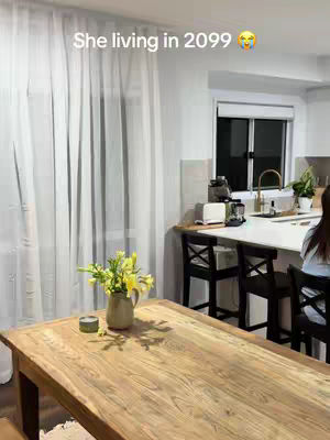 A well-lit interior shot featuring a wooden table with a vase of yellow flowers, and a curtain-covered window in the background. The image suggests a comfortable living space, with a kitchen bar area visible in the background.