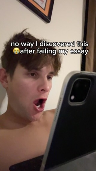 A young man with short brown hair is shown in a close-up, reacting with surprise and his mouth agape as he looks at a phone. The background is a blurred interior with a framed picture visible, the lighting appears to be from a lamp. The color palette is warm, with skin tones prominent and muted background hues. The composition focuses mainly on the man's face, capturing his emotion.