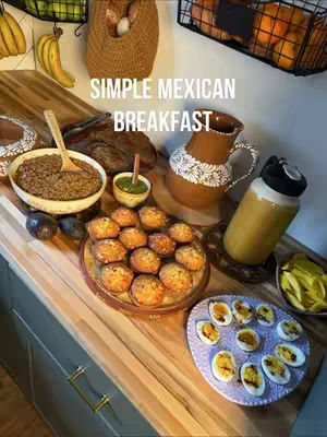The image showcases a kitchen counter filled with various breakfast items. A bowl of cooked food is centrally positioned with a wooden spoon in it. Several baked goods are on a decorative tray, and a plate with sliced boiled eggs is also visible. A brown pitcher, a large water bottle, and various fresh ingredients like avocados and fruits such as bananas and oranges are arranged around the dishes. The overall composition suggests a homemade, visually appealing meal.