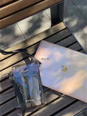 The image shows a laptop and a clear tote bag on a wooden surface, likely a bench. The laptop is open, with a light pink/purple gradient coloring seen on the back. A clear tote bag is positioned to the left, with a black strap visible. The background consists of a darker wooden surface with a few shadows, suggesting daytime sunlight.