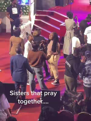 An interior shot of an event showing several people, mostly women, in a church setting. A group of women are embracing in the middle with other people around, and stairs in the background. The color scheme is warm, with red and brown tones.