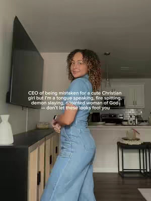 A woman with curly hair stands in profile, looking back at the camera with a slight smile. She wears a denim jumpsuit, and the background shows a home interior with a large TV and a kitchen area visible in the distance. Text overlayed on the image provides additional context.