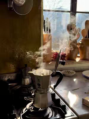 A well-lit, intimate shot of a kitchen scene capturing the preparation of Italian coffee. The foreground features a steaming stovetop espresso maker, with steam rising dramatically. The background includes a window with sunlight streaming through, illuminating various kitchen items like knives in a wooden block and decorative objects. The scene exudes warmth and a sense of home.