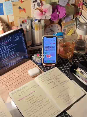 A well-lit and organized desk scene with a focus on study materials and tech devices. In the foreground is an open notebook with handwritten notes, a tablet with its screen displaying what looks like code, and a smartphone on a stand. A pair of AirPods rests near the notebook. In the background, there are various stationery items, notes, and a clock, creating a sense of a productive and aesthetically pleasing workspace. The overall color scheme is bright, clean with neutral and pastel colors.