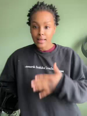 A woman with short, dark hair stands facing the camera, her eyes open and mouth slightly forming a speaking position. She wears a black sweater over a red t-shirt, with green background. She is positioned in front of a wall, and her right arm is extended towards the direction of the camera, while her left arm is near her body. The lighting is bright and evenly distributed, highlighting her face and the surrounding environment.