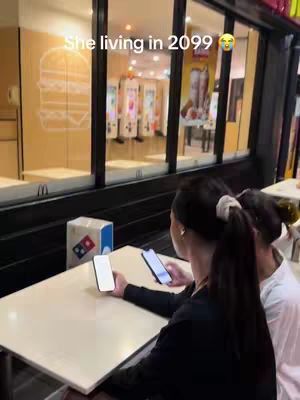 A woman is seated at a cafe table, holding two smartphones. She is facing away from the camera, her dark hair tied back in a ponytail. The background includes the windows of a restaurant, with a blurred view of the interior that seem to be a fast food restaurant, and a Domino's pizza box on the table. A text overlay 'She living in 2099' appears at the top of the image.