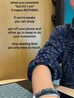 The image shows a close-up of a person's arm with a dark blue sweater and a beaded bracelet. The person's arm is resting on a keyboard, which implies they are working or studying. Text overlaid on the image reads "when you comment 'but it's 1 am' it means NOTHING, if you're awake you can study, get off your phone and either go to sleep or do your homework, stop wasting time you only have so much." The background is a wooden surface.