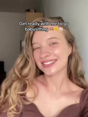 A young woman with blonde hair smiles at the camera. She has a natural makeup look and is wearing a brown top. The image is framed from the chest up. Text overlay reads, "Get ready with me to go babysitting".