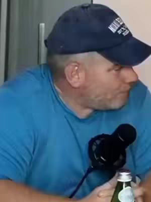 A man wearing a blue cap and shirt is seated, facing slightly to the side. A microphone is positioned in front of him. A bottle of water is being held in his hand. The lighting appears to be natural.