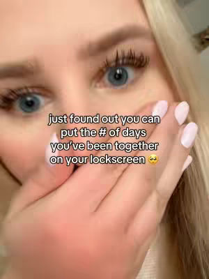 A close-up shot features a fair-skinned woman with long blonde hair, partially covering her face. Her blue eyes are wide and accented with dark eyelashes. A hand with light pink, manicured nails covers her mouth and the lower portion of her face, partially obscuring her lips. The background is blurred, but a wall or surface is visible.